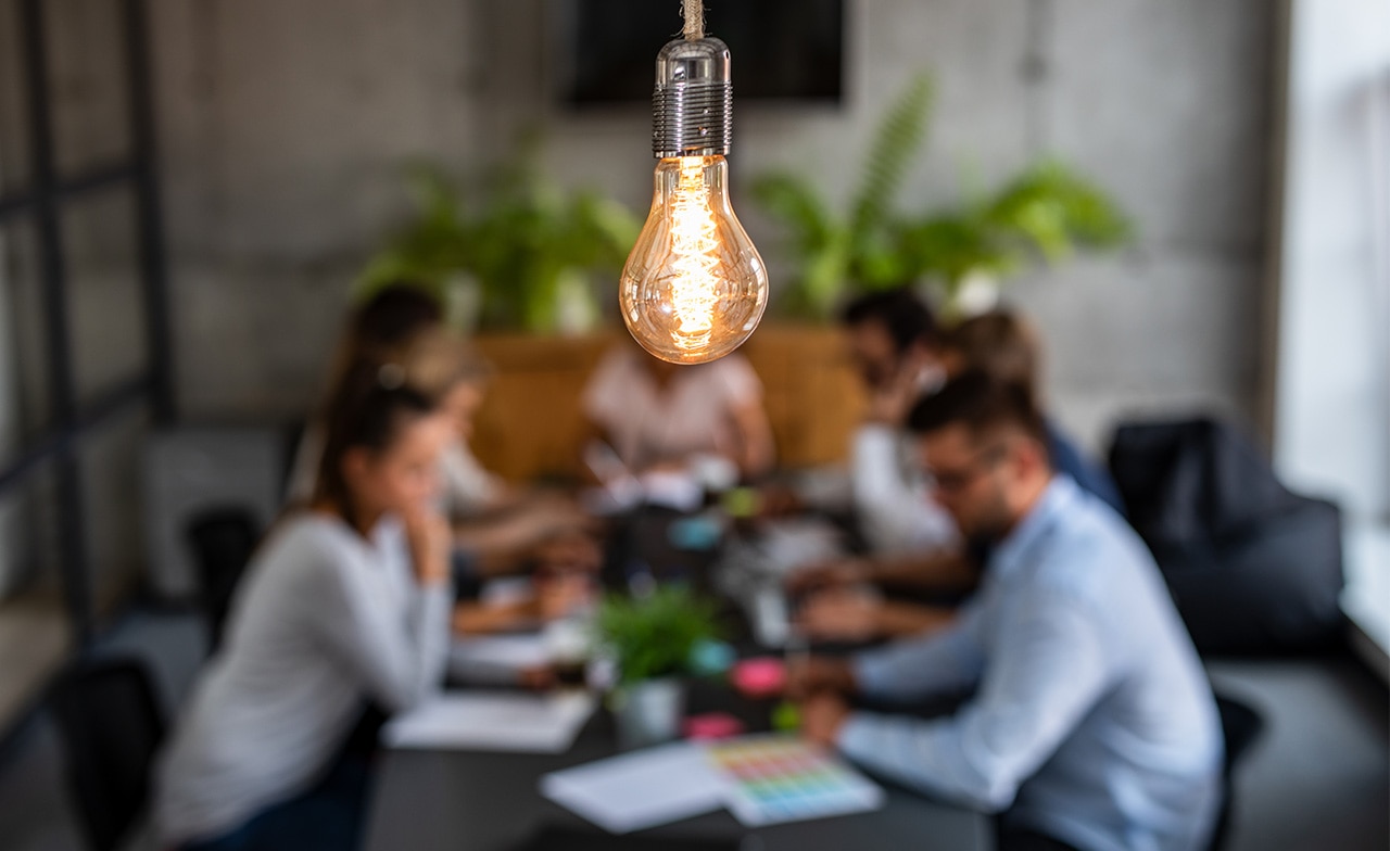 Young creative business people meeting in an office with a lightbulb in the foreground shining overhead Young creative business people meeting in an office with a lightbulb in the foreground shining overhead
