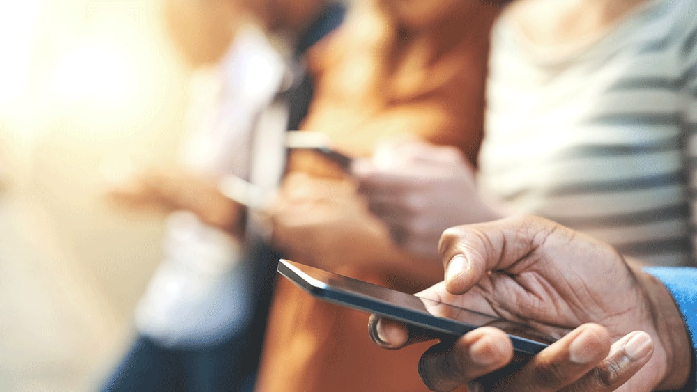Man’s hand holding a mobile phone with other people in the background Man’s hand holding a mobile phone with other people in the background