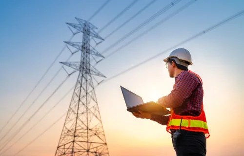 Engineer in hi-vis vest using a laptop near power lines at sunset