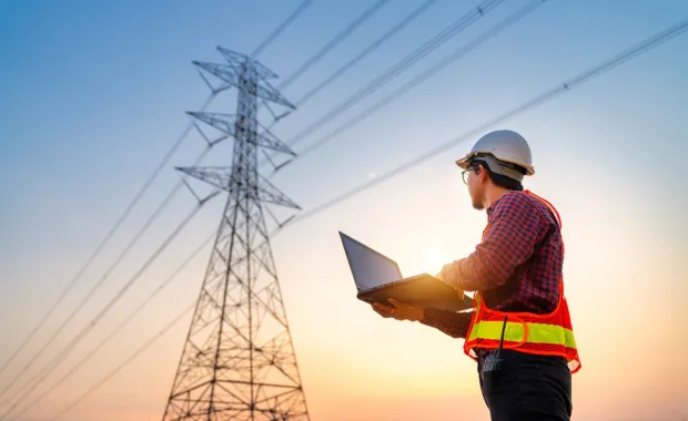 Engineer in hi-vis vest using a laptop near power lines at sunset