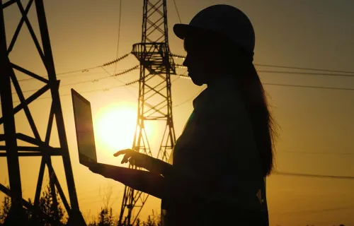 engineer works on laptop by electrical towers a silhouetted electrical engineer records data on a laptop next to electrical towers at sunset